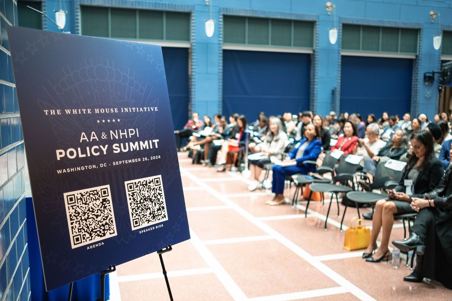 AA & NHPI Policy Summit sign with QR code in foreground, attendees seated in background at White House event.