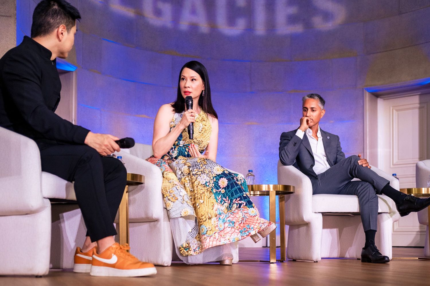 Three panelists discuss on stage at a Legacies event, with a woman in floral dress speaking into a microphone.