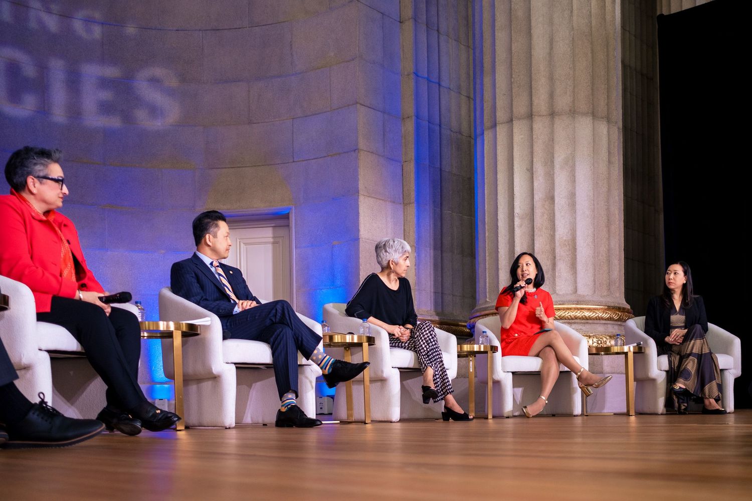 Five panelists seated on stage in elegant white chairs during a formal conference discussion under blue ambient lighting.