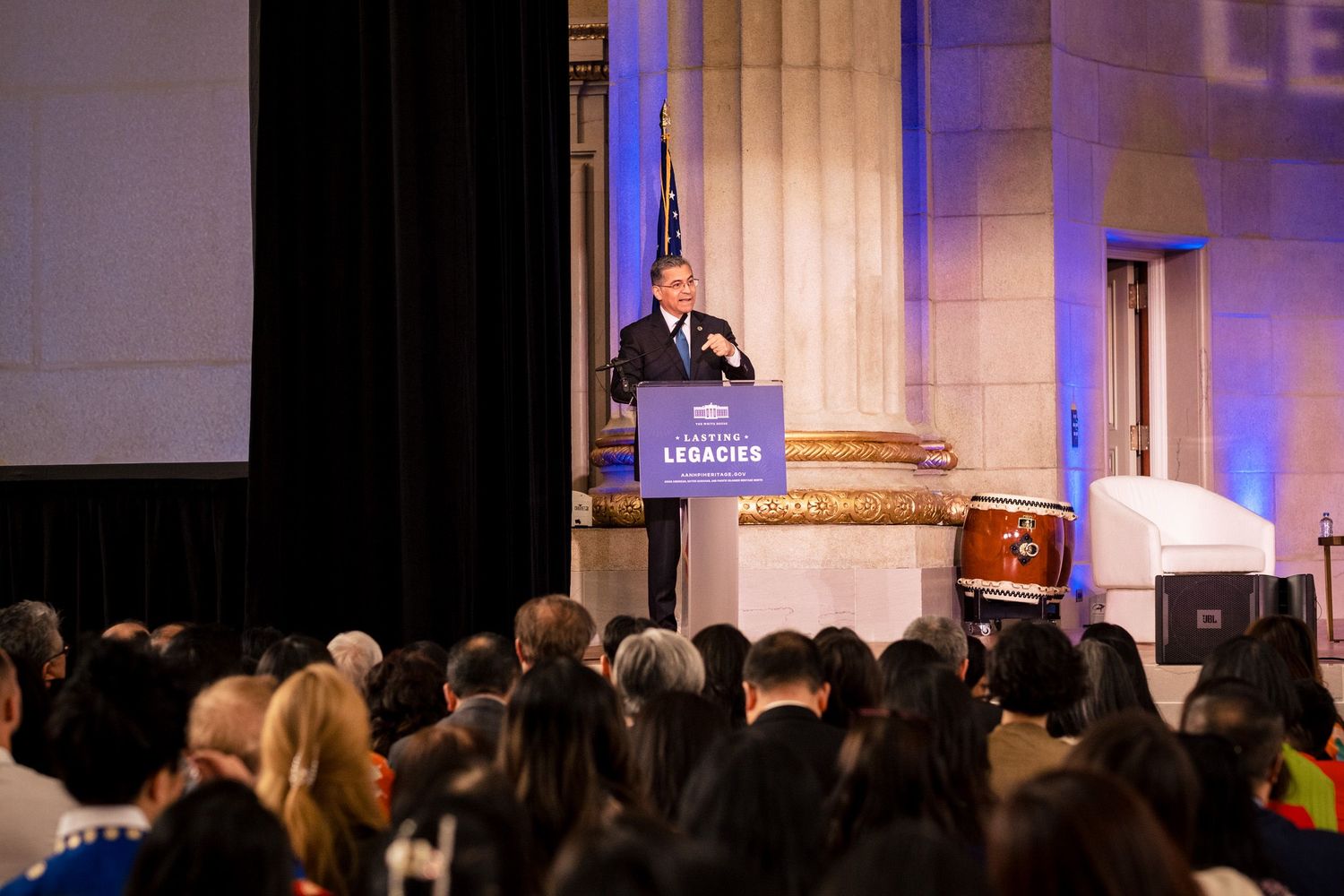 Speaker at podium addresses large crowd at a Legacies event in grand hall with blue decor.