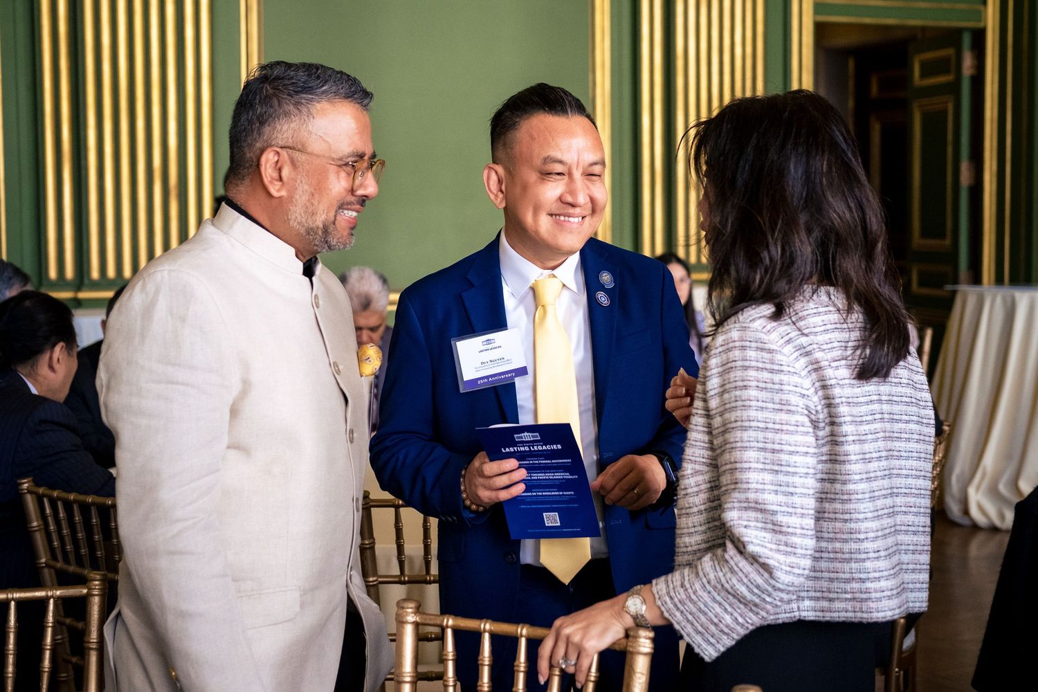 Three professionals networking at a formal event, man in blue suit holding award plaque in elegant green room.