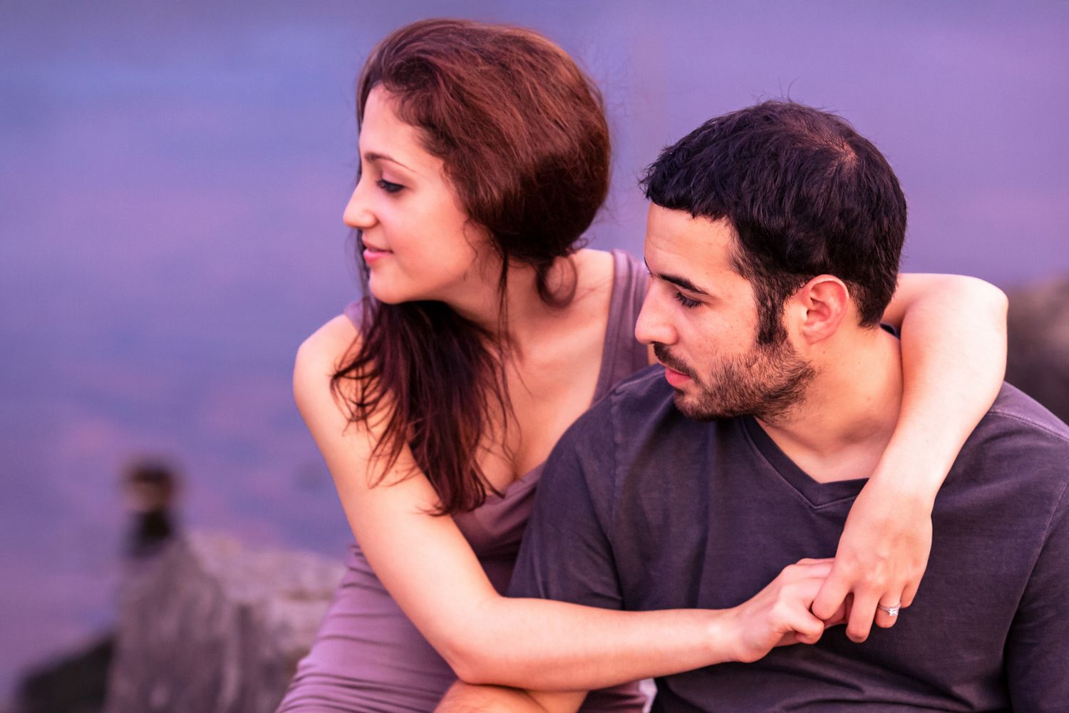 Couple sitting close together by water at dusk, woman embracing man from behind.