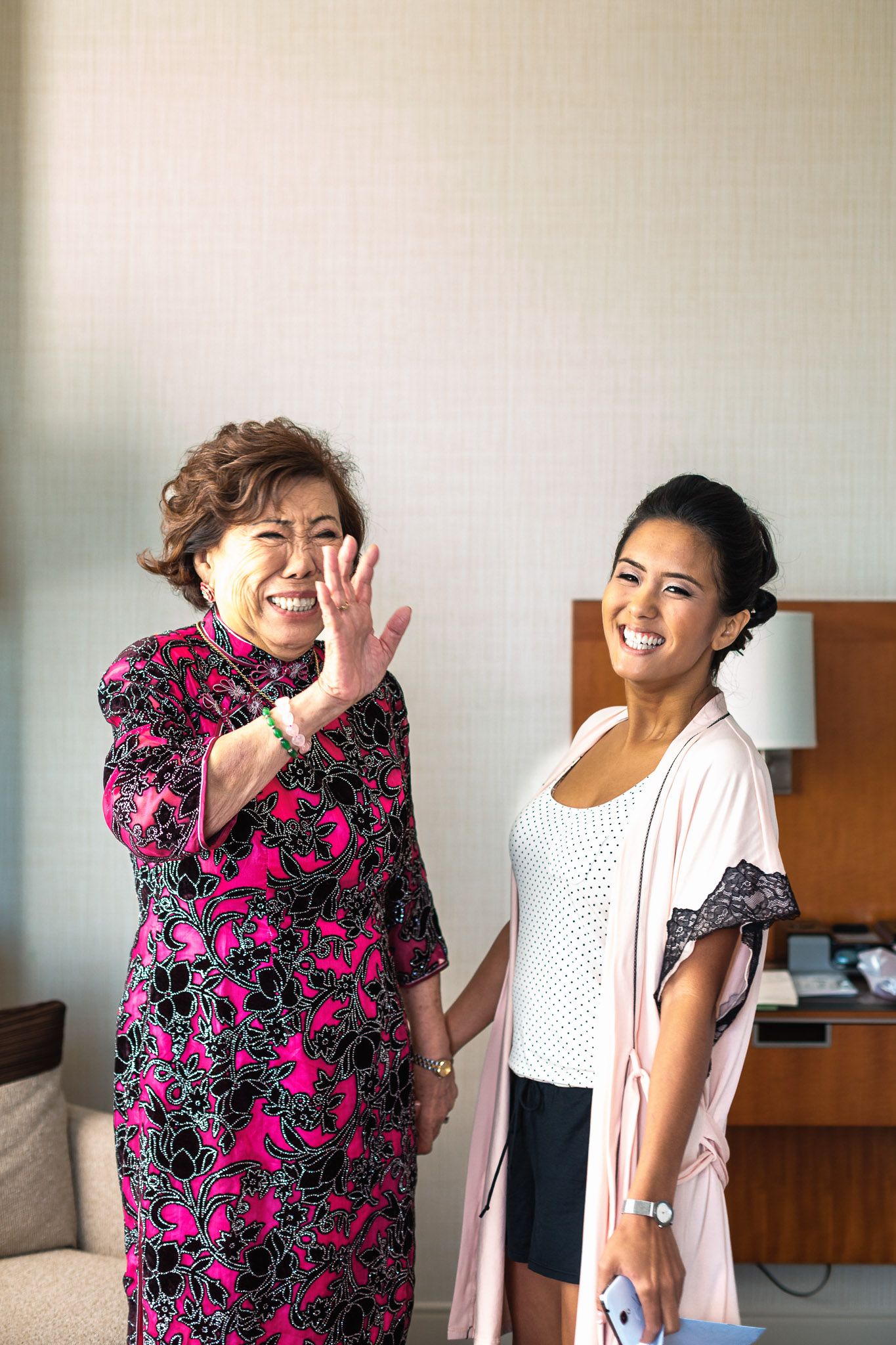 Older woman in pink floral dress waves beside smiling younger woman in a hotel room.