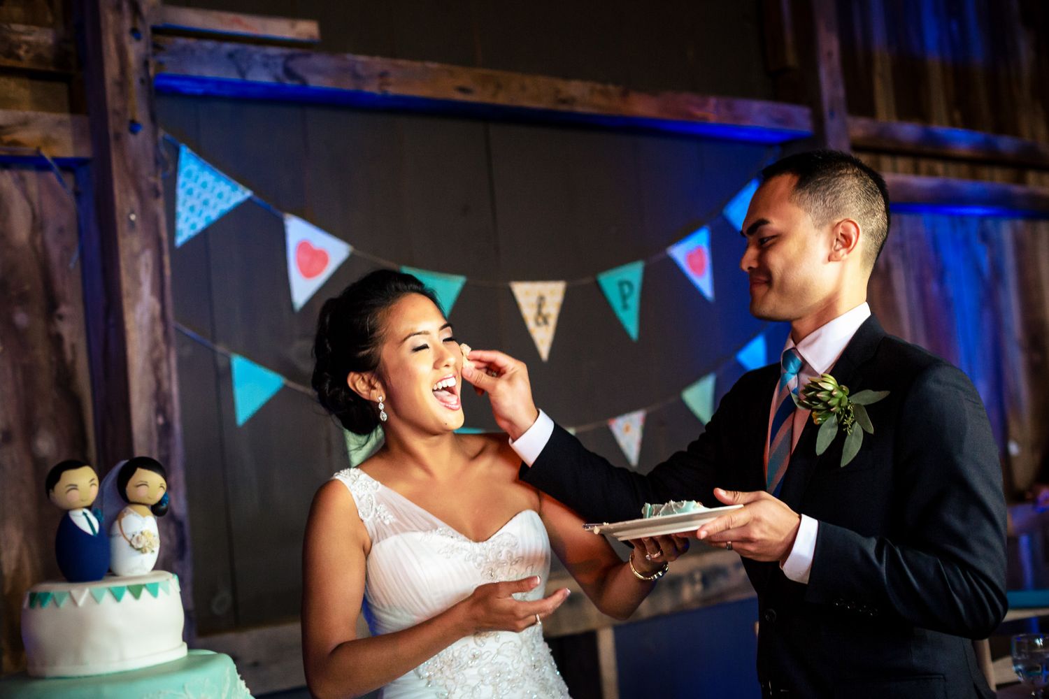 Groom feeds wedding cake to his laughing bride near a decorated cake in a rustic barn reception.
