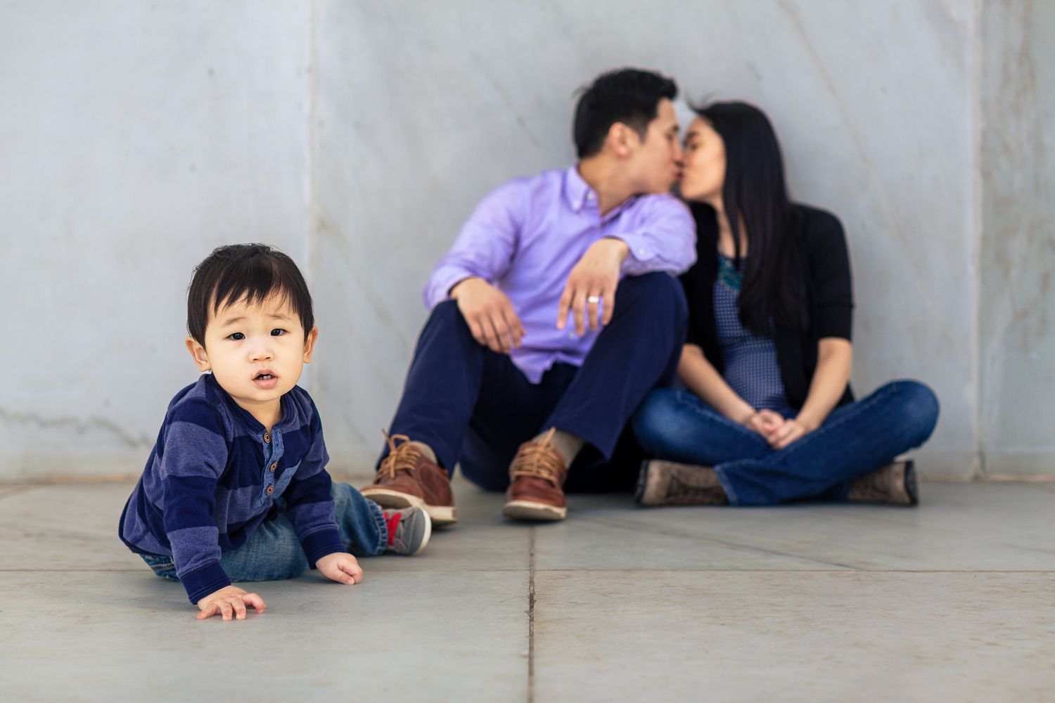 Asian toddler crawls on floor while parents share a kiss in the background against a wall.