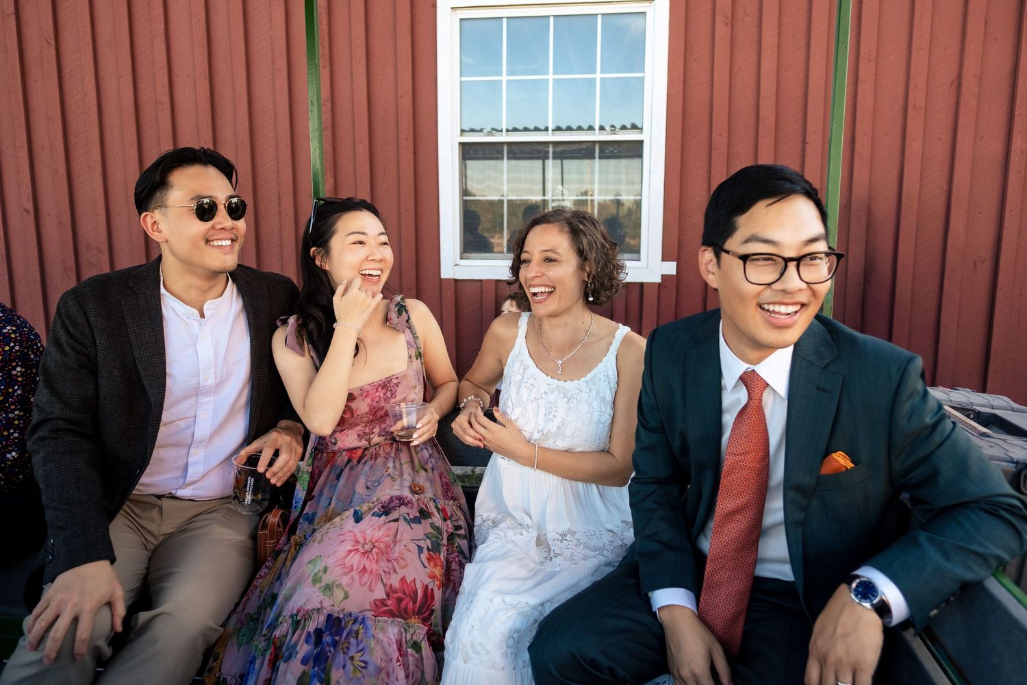 Two couples laughing and sitting together outside a red barn at a wedding celebration.