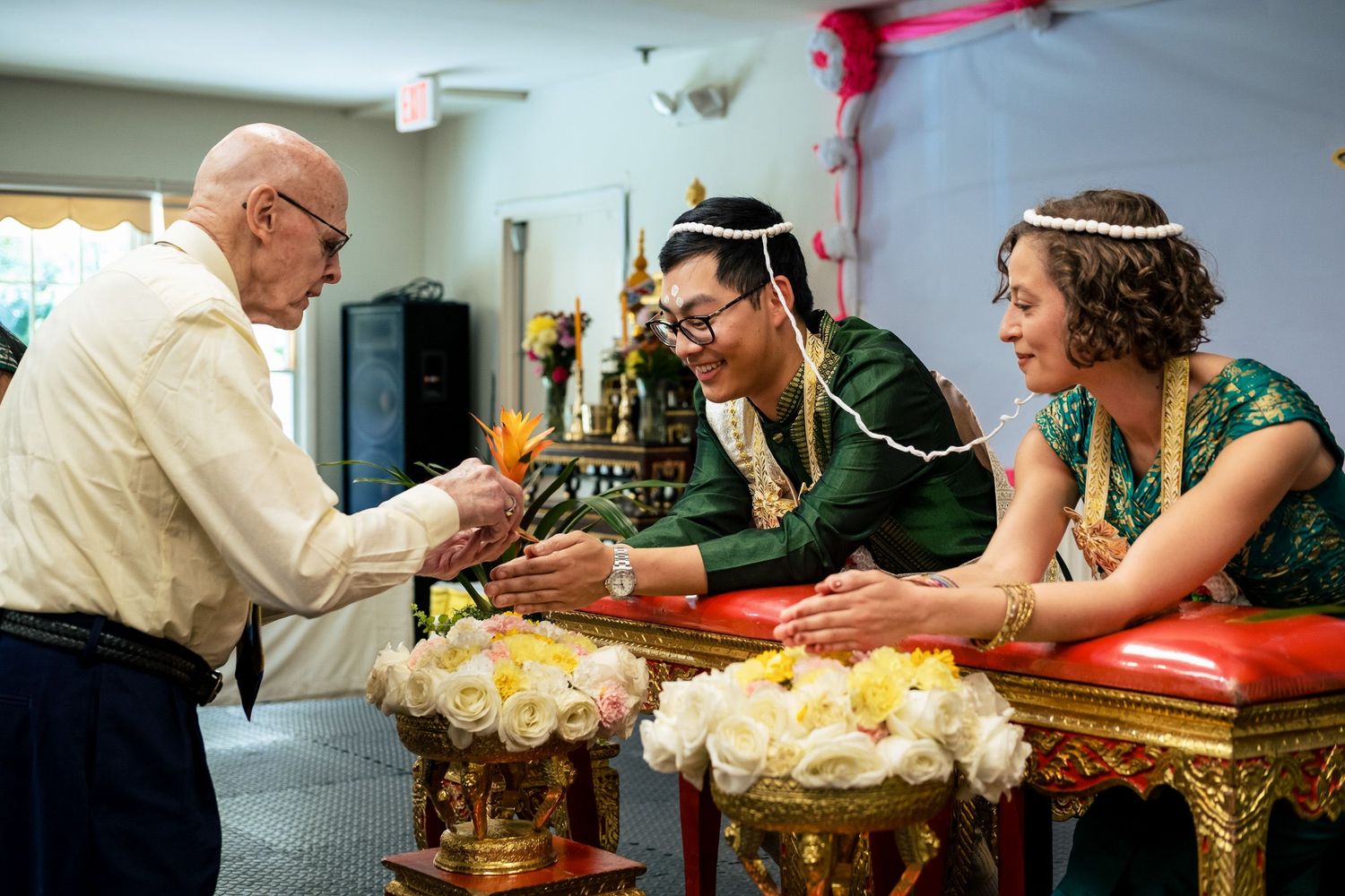Elderly man offering blessings to couple in traditional Thai wedding garments at ornate ceremony table.