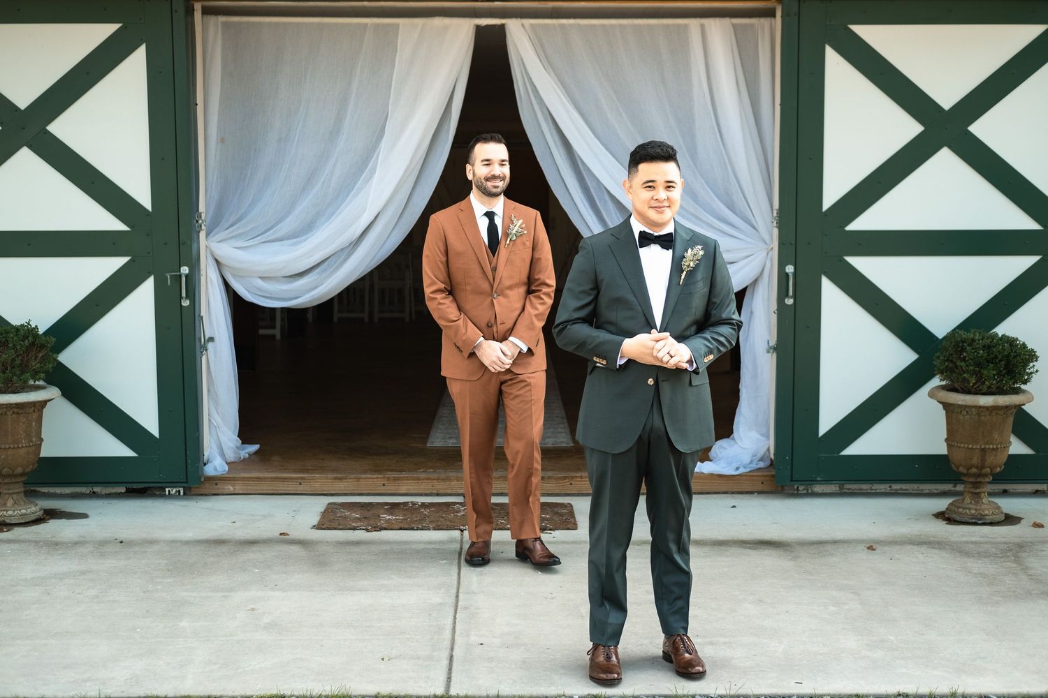 Two grooms in contrasting brown and black tuxedos standing together at barn venue entrance.