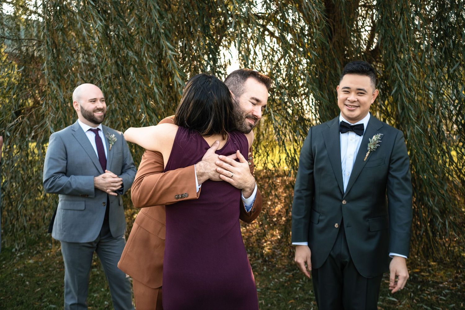 Groom in brown suit hugs female officiant in purple dress while other groom in tuxedo watches and smiles.