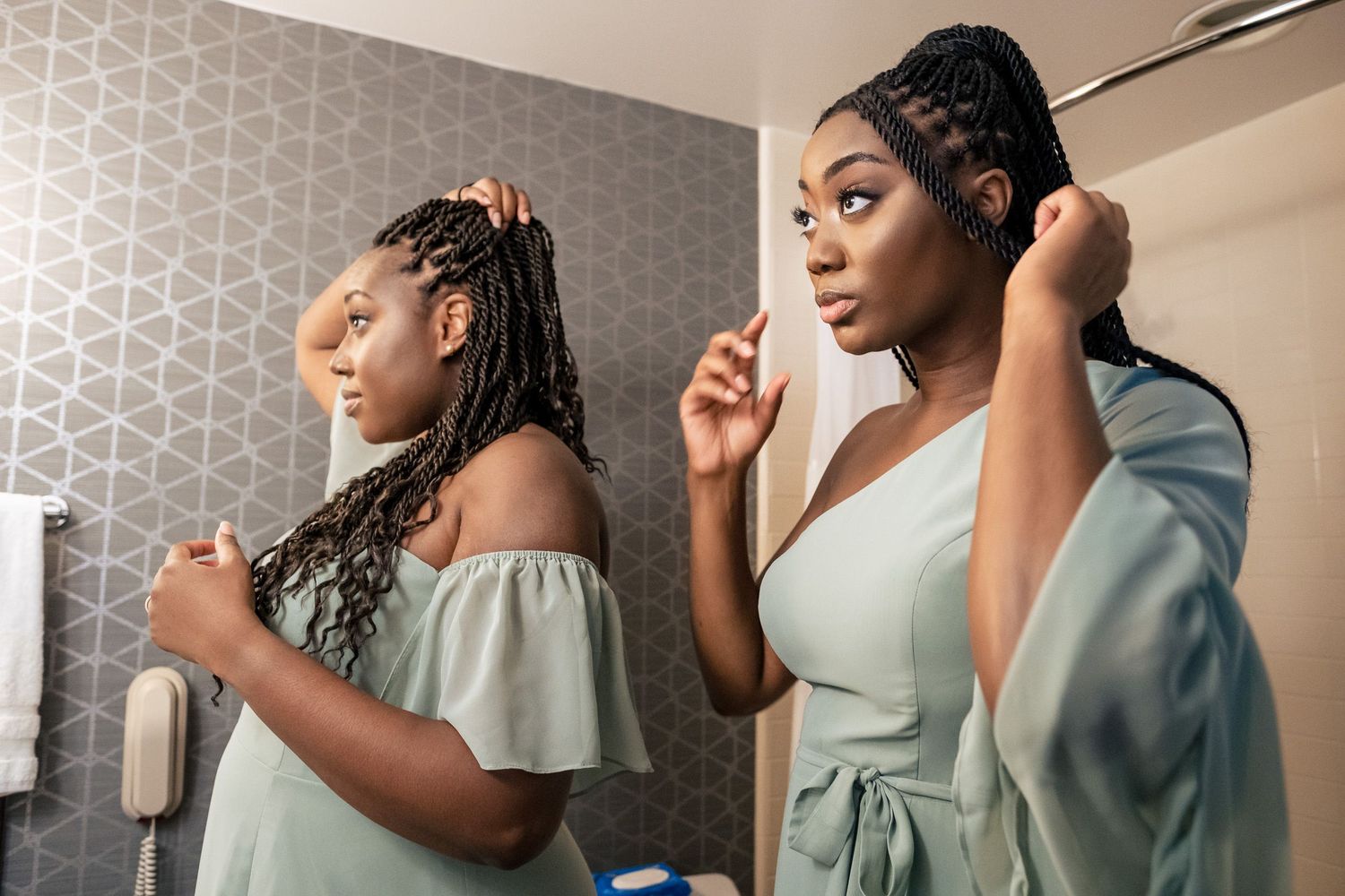 Two Black women in sage green dresses style braided hair while getting ready for a wedding.