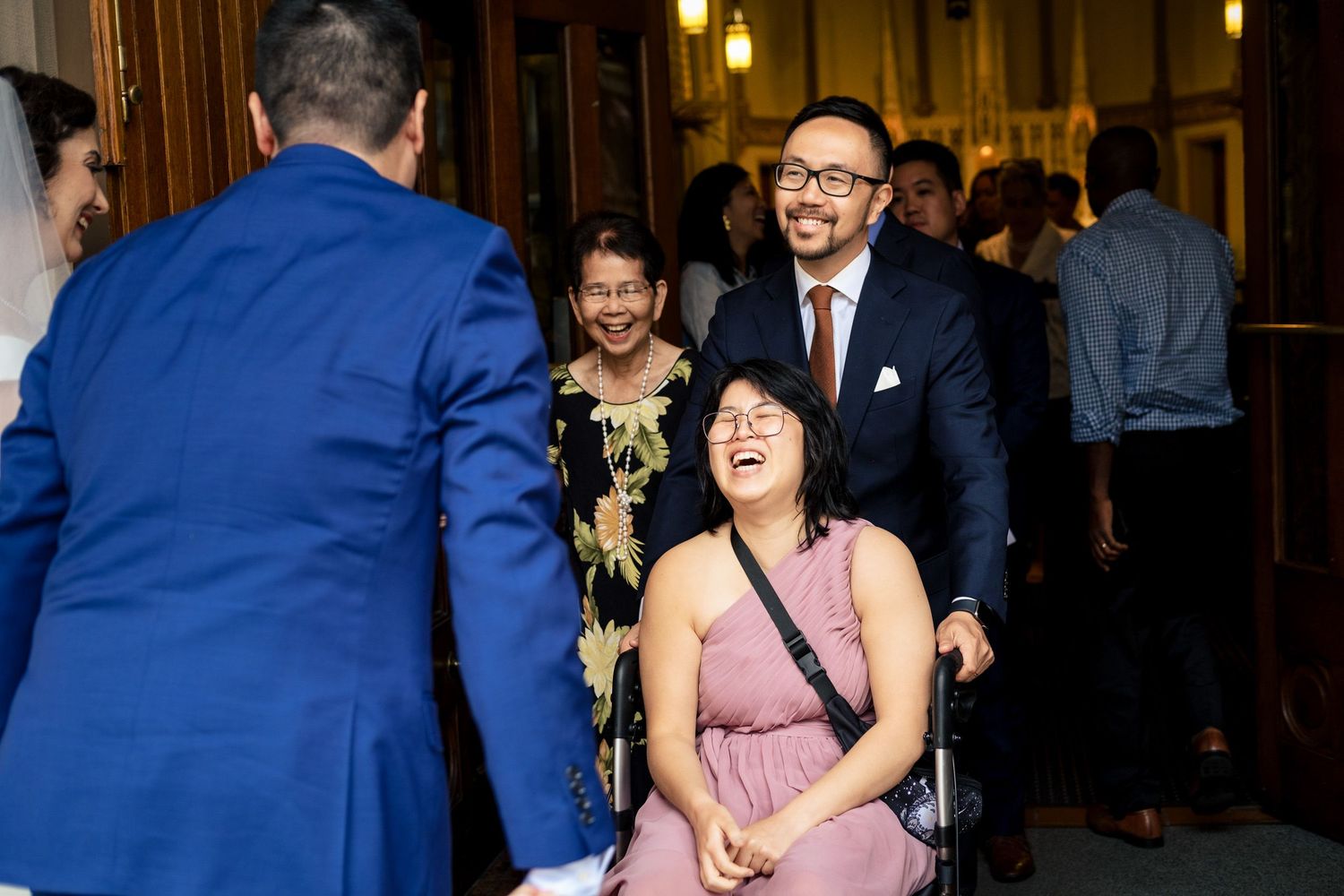 Laughing wedding guest in wheelchair greeted by groom in blue suit among smiling guests at elegant venue entrance.