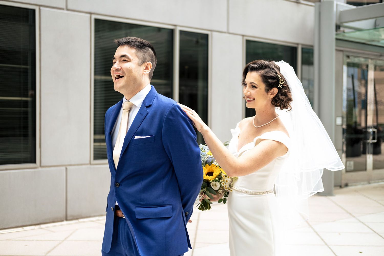 Bride in white gown with veil taps groom on shoulder during outdoor first look wedding moment.