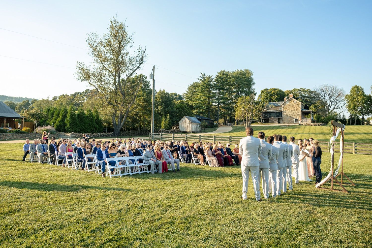 Wide view of guests seated in white chairs for an outdoor wedding ceremony on a sunny day at a farm venue.