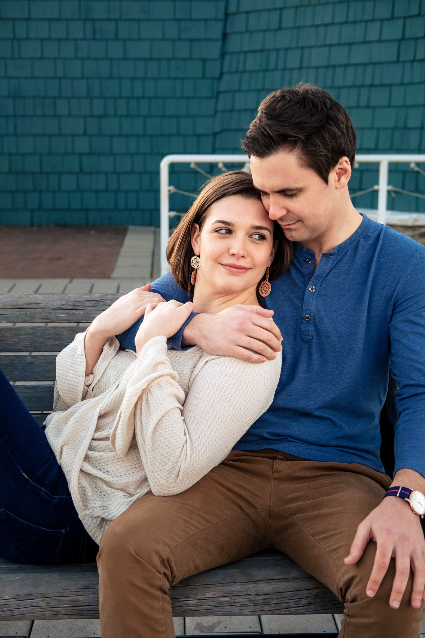 Couple sitting on wooden steps, man embracing woman from behind, both smiling softly outdoors.