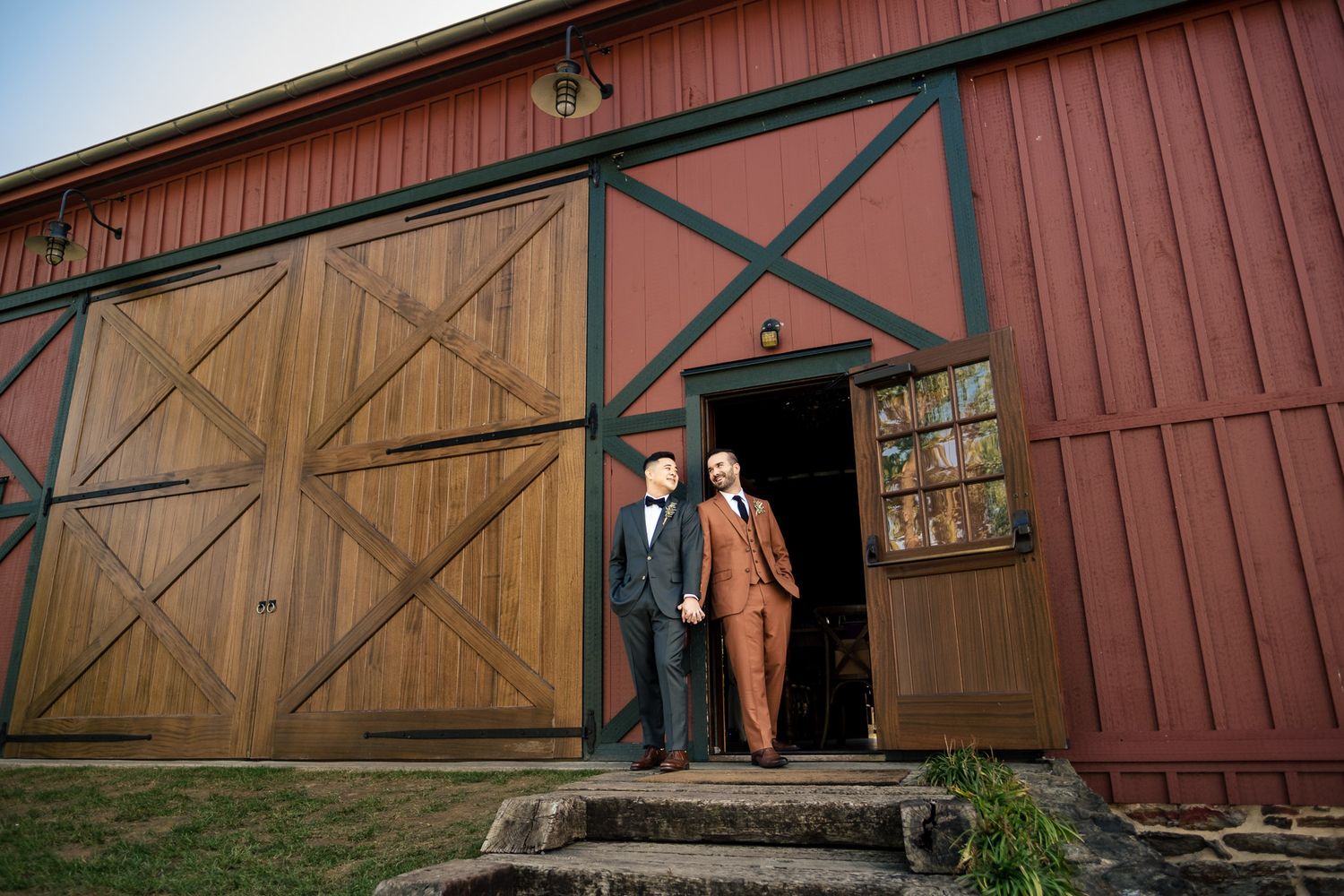 Two grooms in suits stand in the doorway of a rustic red barn wedding venue.
