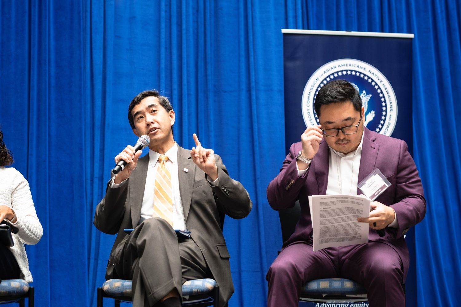 Two Asian men on stage at a formal panel discussion, one speaking into a microphone while holding up a finger.