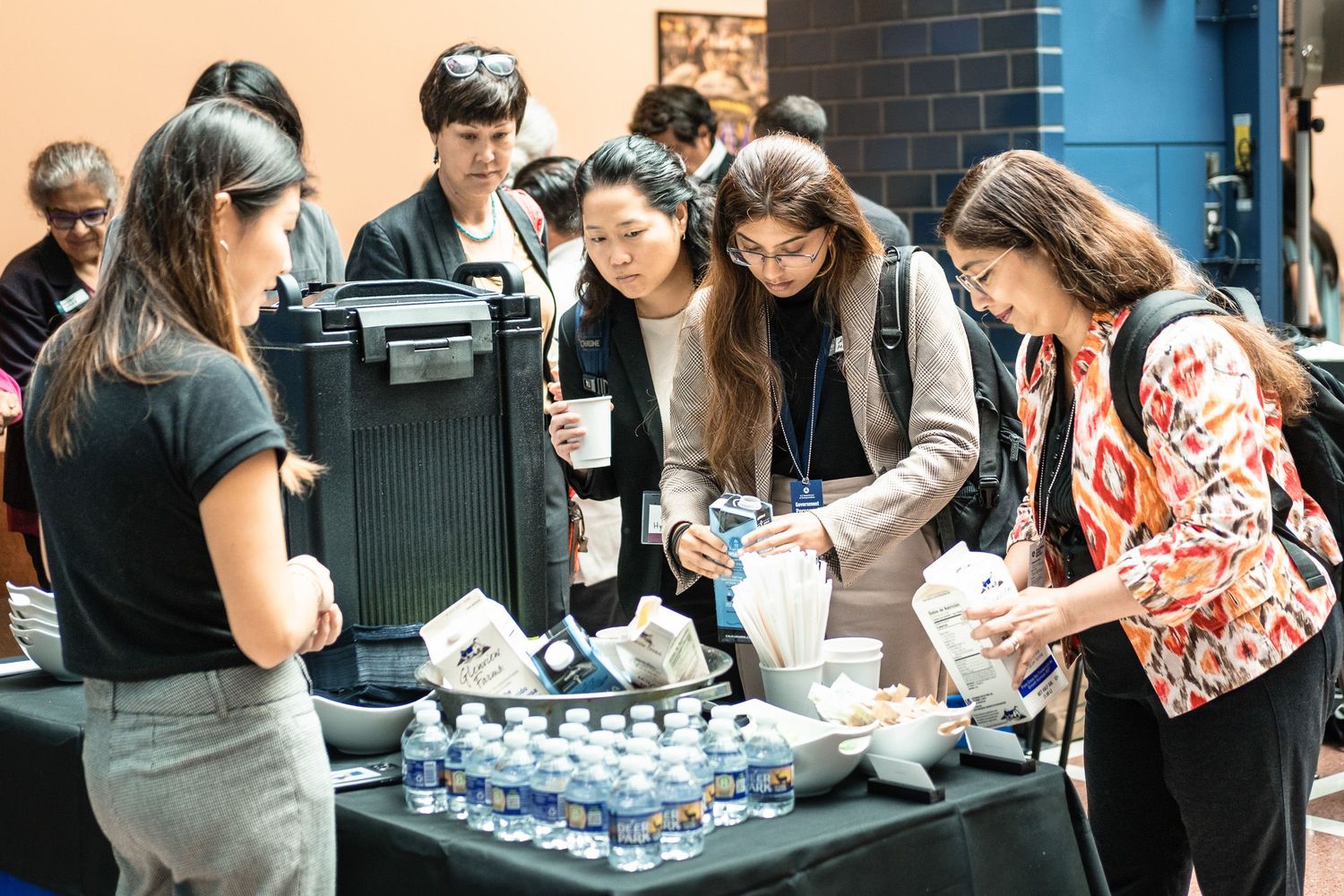 People gathering around a catering table with food and water bottles at an indoor event or conference.