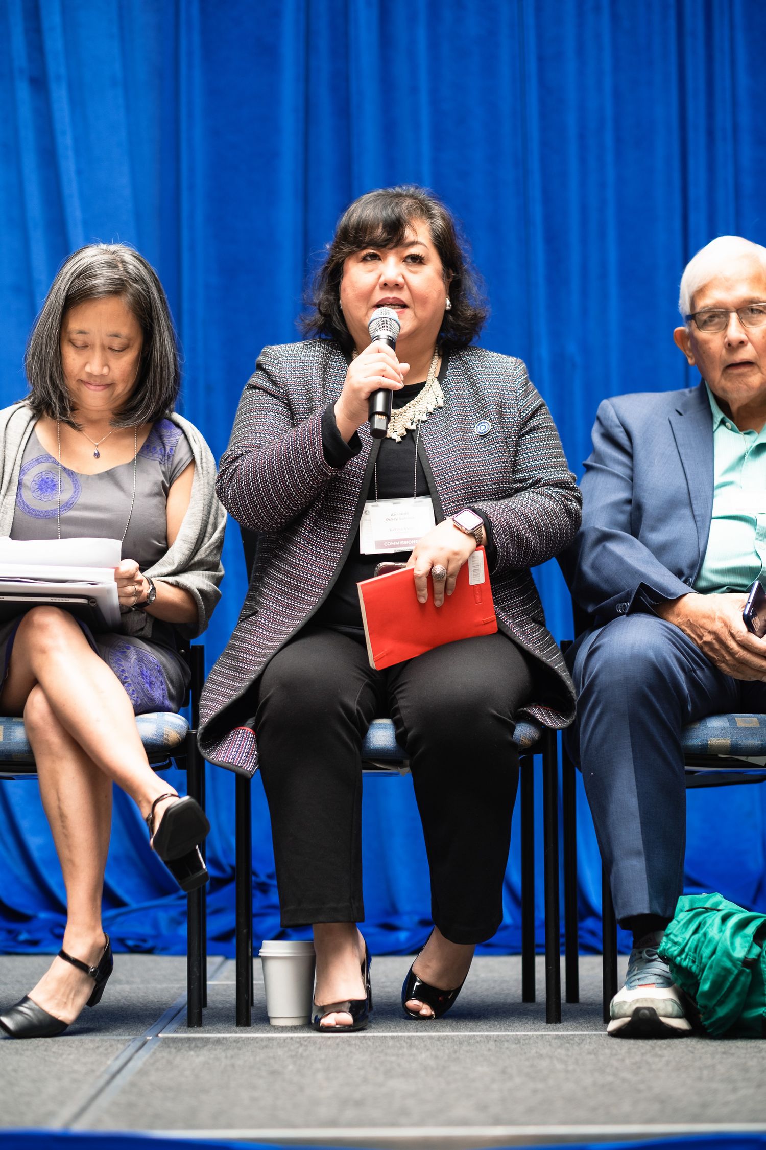Woman speaking into microphone at panel discussion, flanked by two seated attendees against blue curtain backdrop.