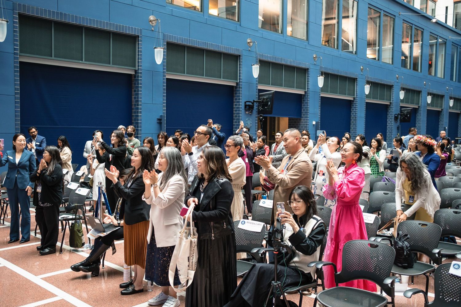 A diverse crowd of attendees applauds at an indoor graduation ceremony outside a blue brick building.
