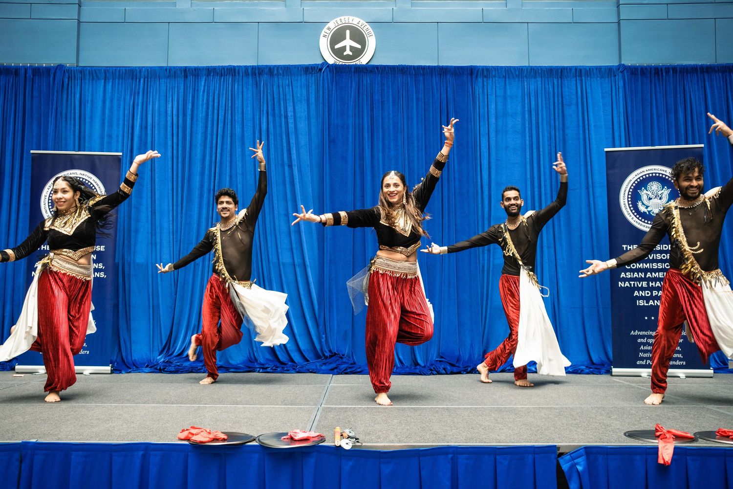 Colorful Bollywood dancers performing on stage with blue curtain backdrop at a cultural event.