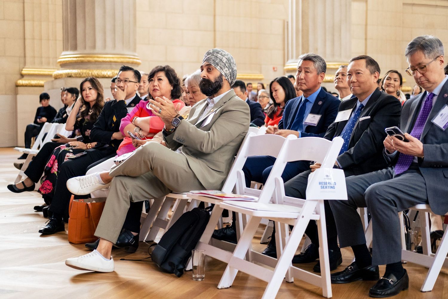 Diverse group of people seated in white chairs at a formal indoor event or ceremony.