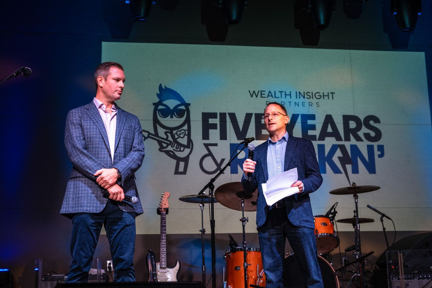 Two men on stage at Wealth Insight Partners Five Years and Rockin' anniversary event with guitars in background.