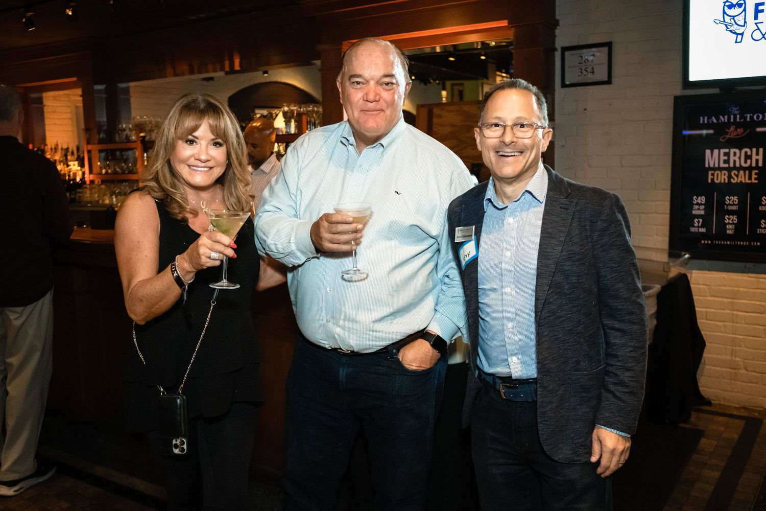 Three smiling adults holding drinks pose together at a casual networking event inside a bar or restaurant.