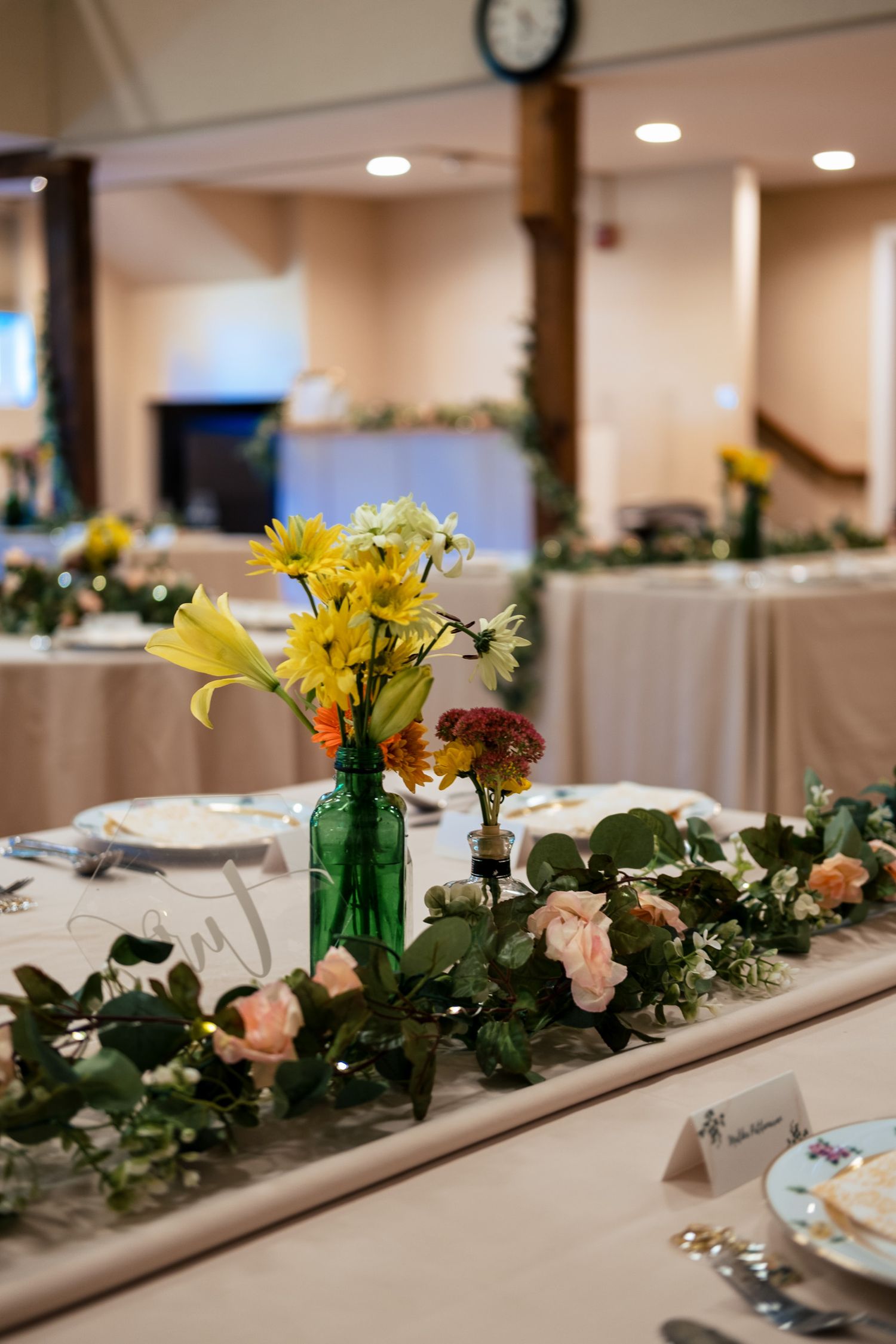 Wedding reception table with green bottle vase holding yellow and orange flowers alongside a pink rose garland runner.