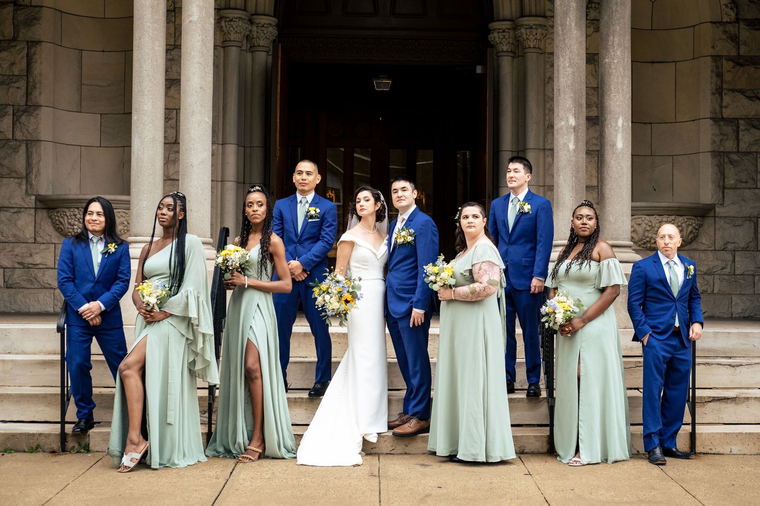 Wedding party in sage green and royal blue poses on stone church steps with bride and groom.