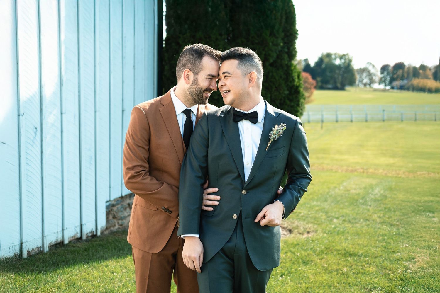 Two grooms in brown and navy suits share a tender moment outdoors at their wedding celebration.