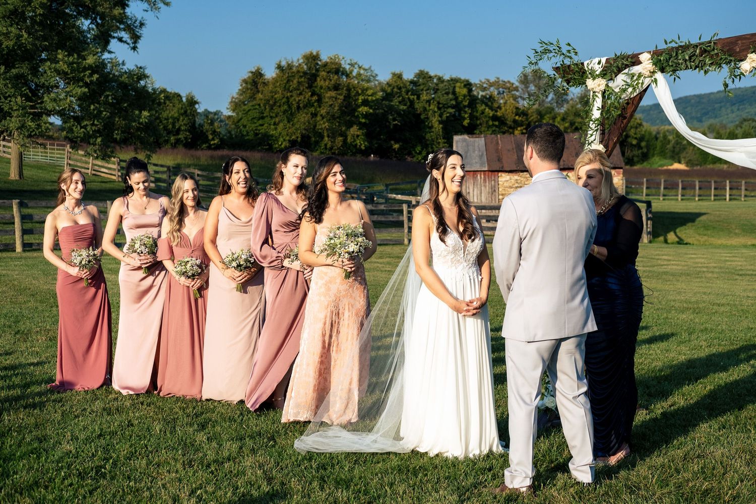 Bride and groom exchange vows before an officiant with bridesmaids in pink dresses standing nearby outdoors.
