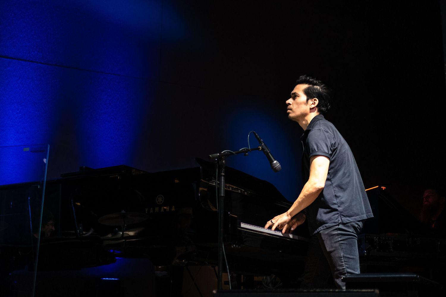 Male musician plays keyboard on stage bathed in blue stage lighting during live concert performance.