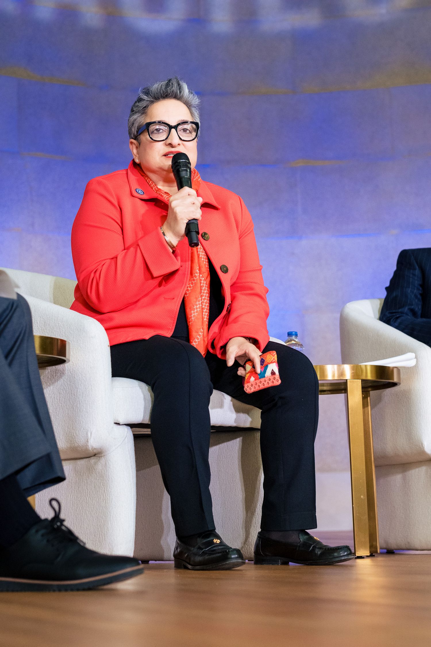Person in red jacket speaking into microphone at panel discussion event on stage.