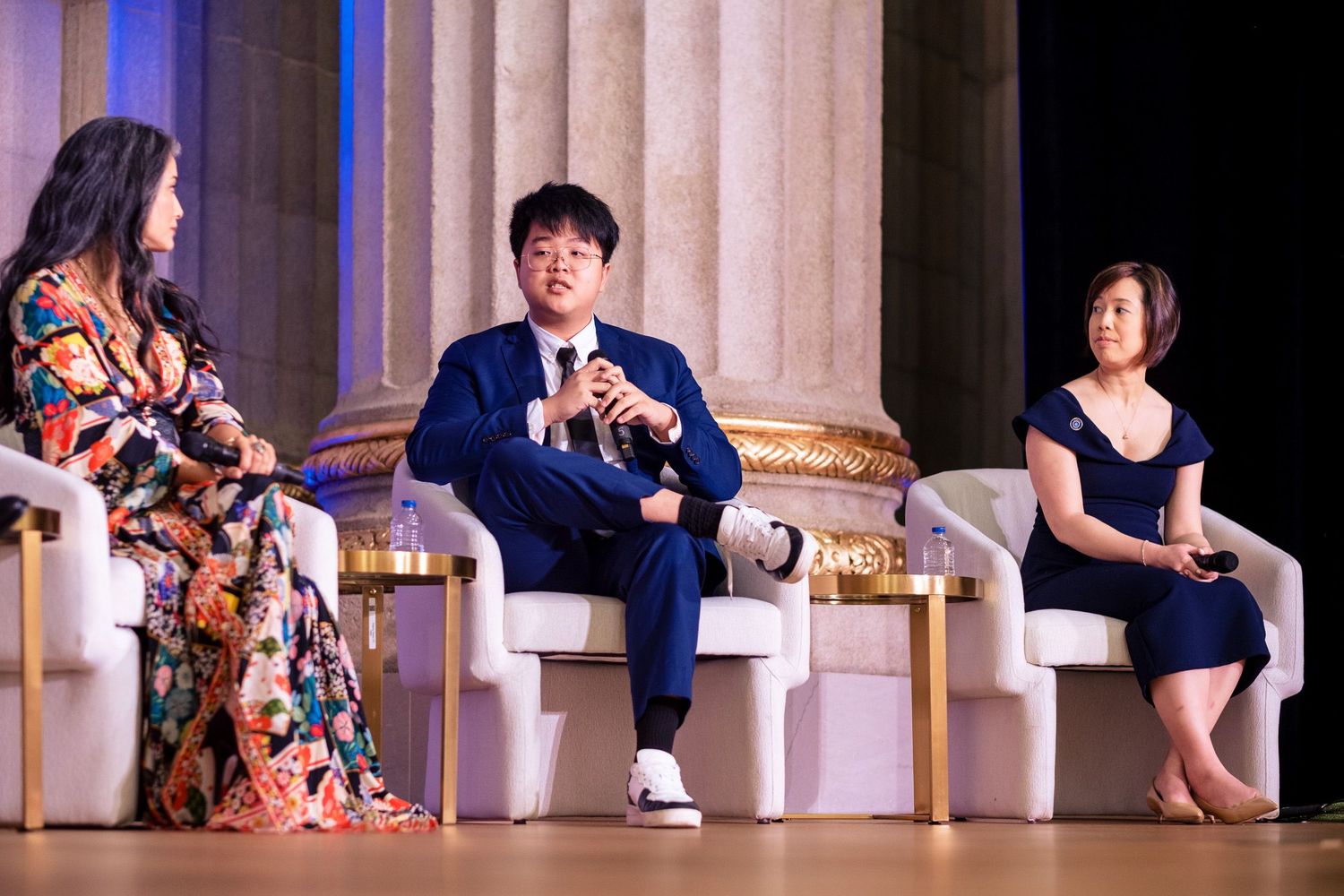 Three panelists seated on stage in elegant chairs during a formal conference event under decorative lighting.