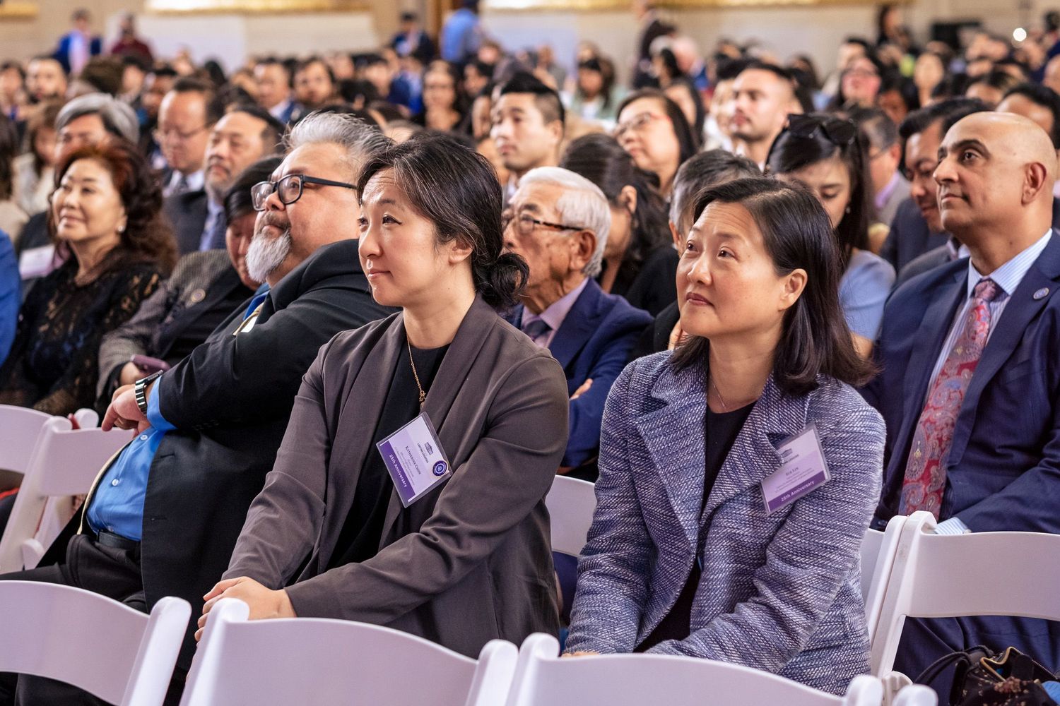 Diverse audience seated at a formal conference event, with attendees wearing name badges and business attire.