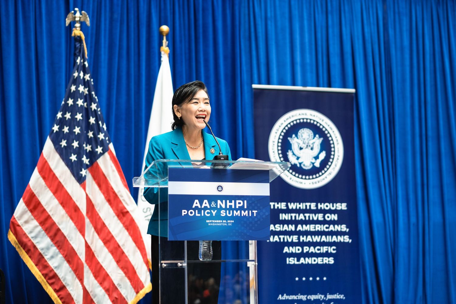 Woman speaking at AA+NHPI Policy Summit podium with US flag and White House Initiative banner backdrop.