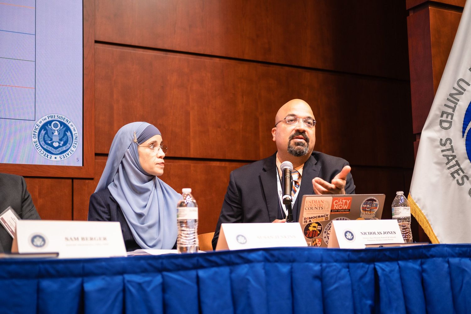 Two panelists speaking at a United States government conference, with a woman in hijab and a bearded man at the table.