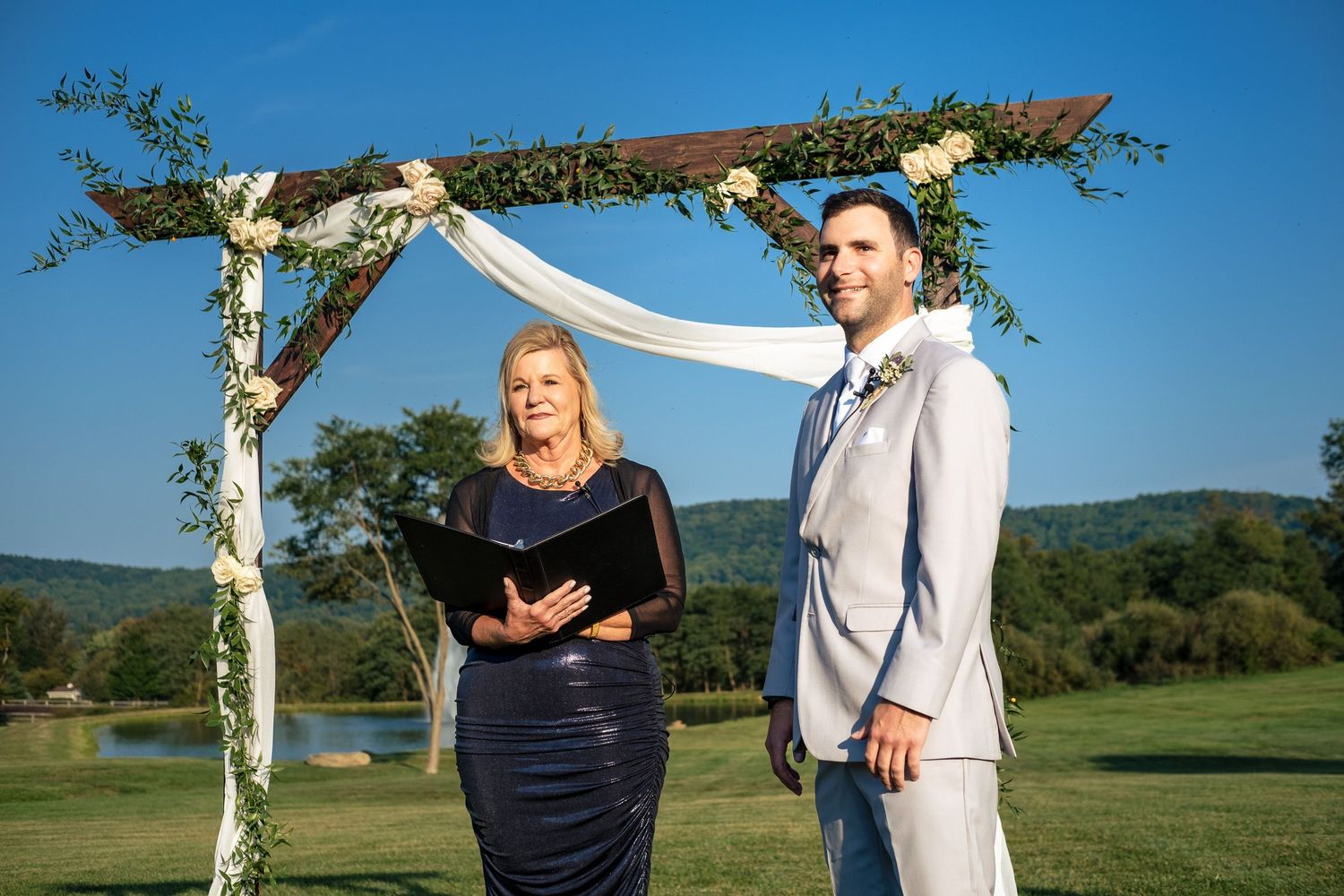 Groom in gray suit stands with officiant under a rustic floral arch during an outdoor wedding ceremony.