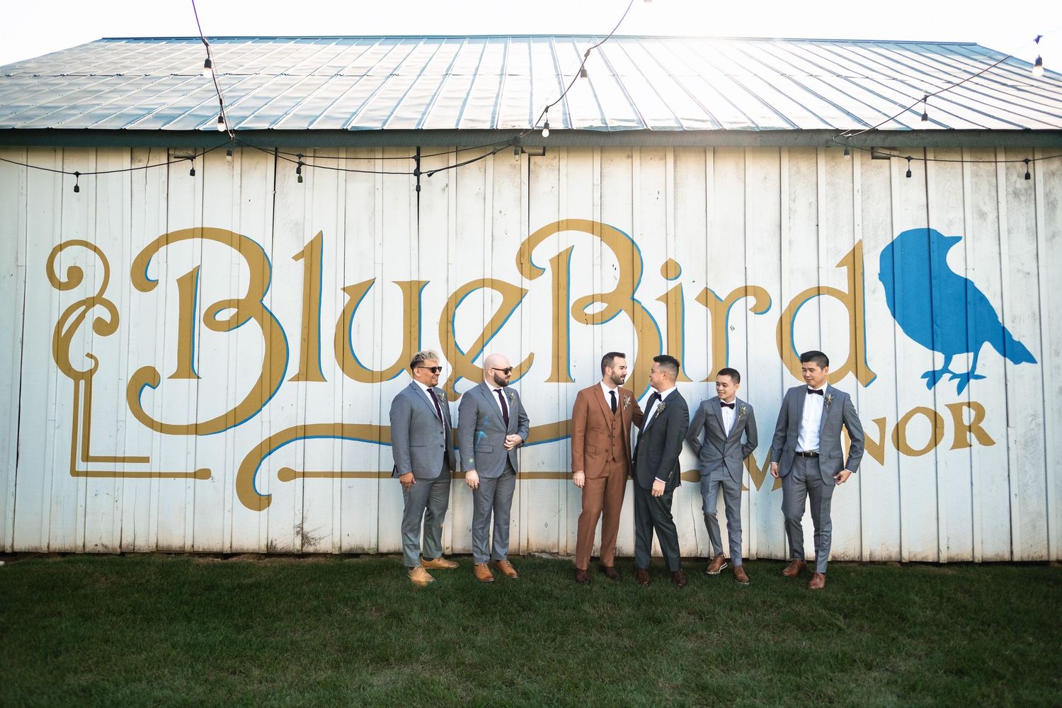 Wedding party of six men pose in suits before Blue Bird Manor barn sign.