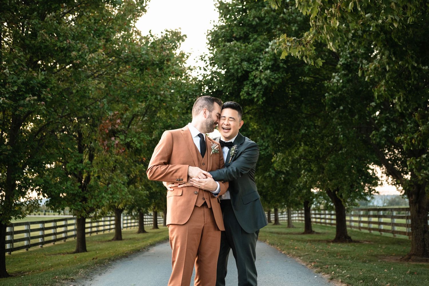Two grooms sharing a joyful laugh together on a scenic tree-lined path at their wedding.