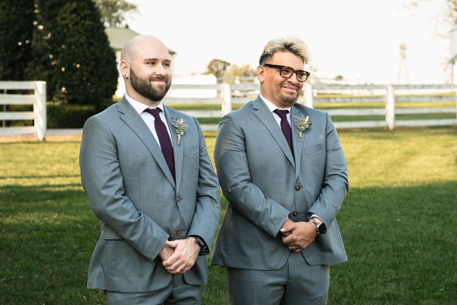 Two groomsmen in matching gray suits with burgundy ties and boutonnieres stand smiling at an outdoor wedding ceremony.