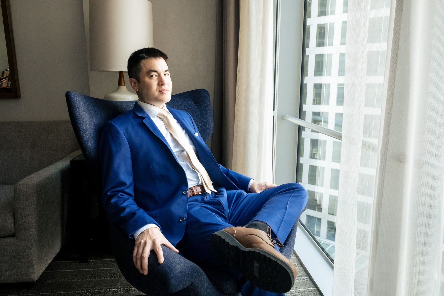 Dapper groom in royal blue suit sitting in chair gazing out hotel window before wedding.