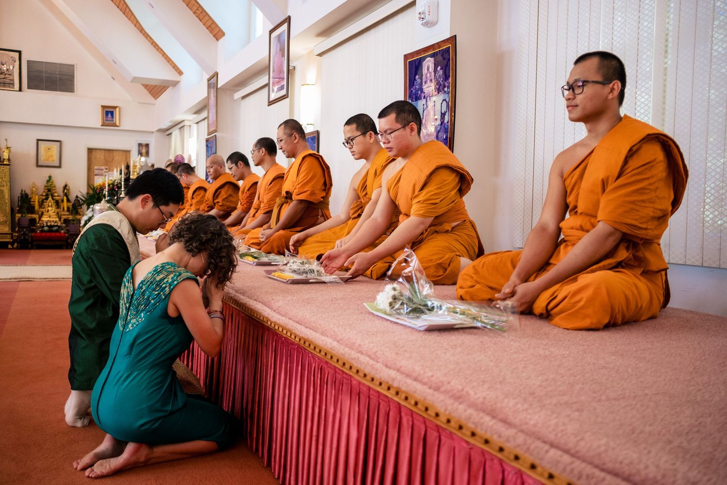 Bride and groom bow before Buddhist monks in orange robes during traditional Thai wedding blessing ceremony.