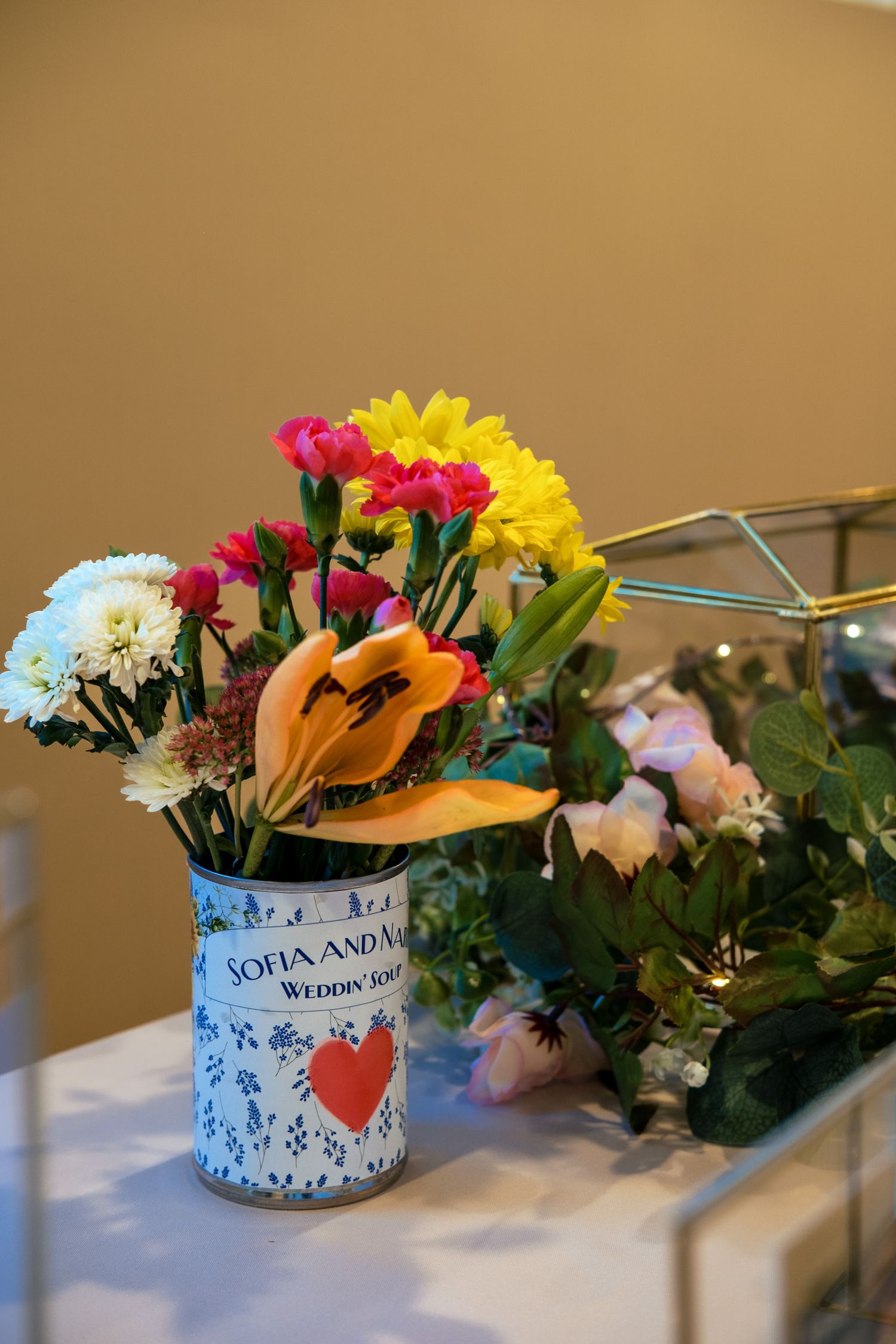 Colorful wedding floral arrangement in a decorative Sofia and Nabin Wedding Soup can vase with pink, yellow, and orange flowers.