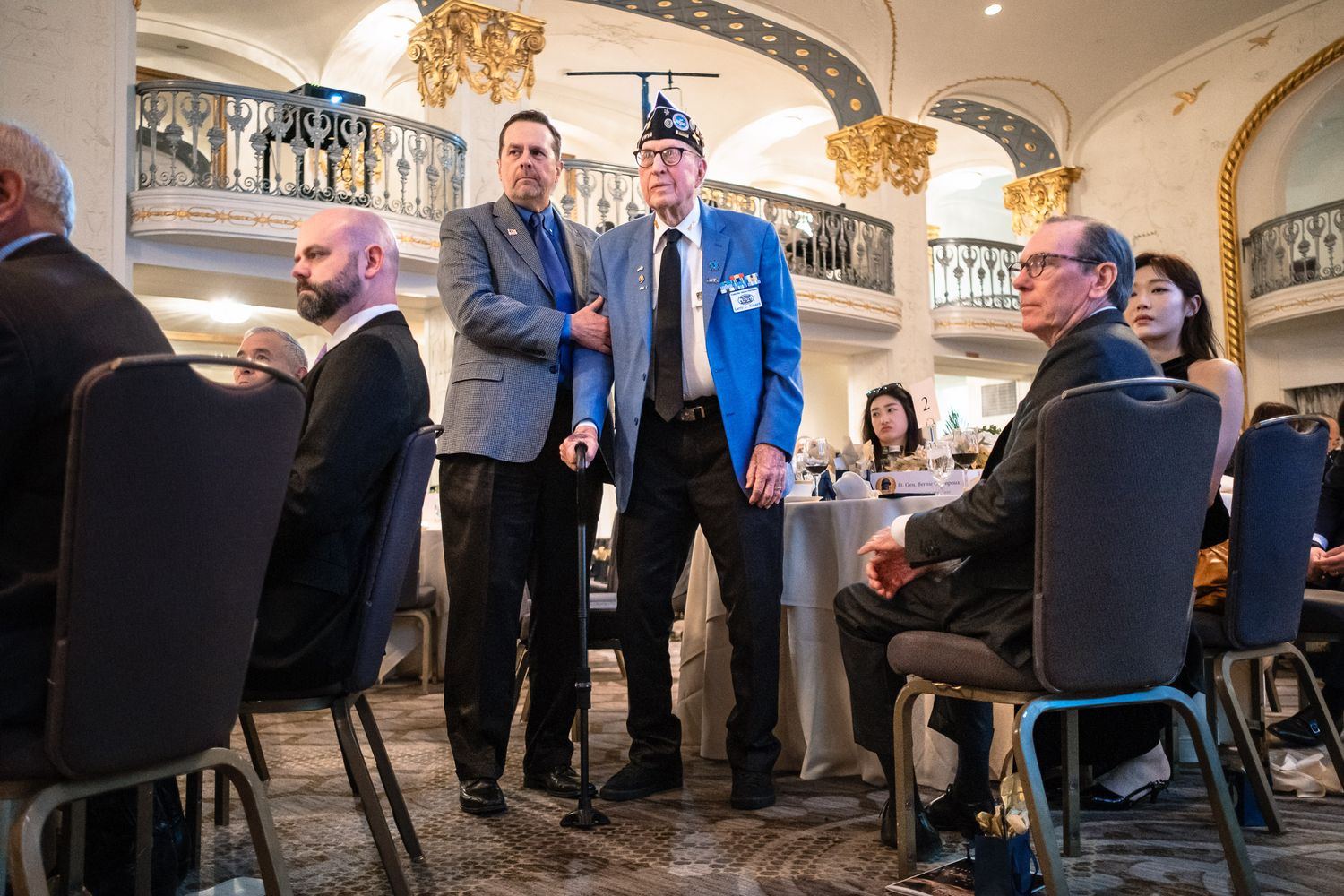 Two men stand and converse at formal banquet event in grand ballroom setting.