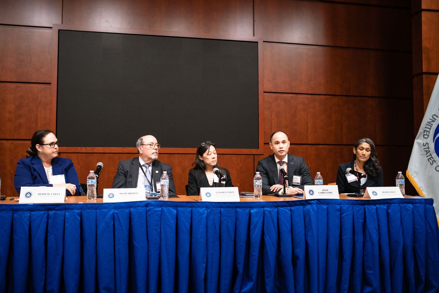 Four professionals seated at a blue-draped conference table engaged in a formal panel discussion event.