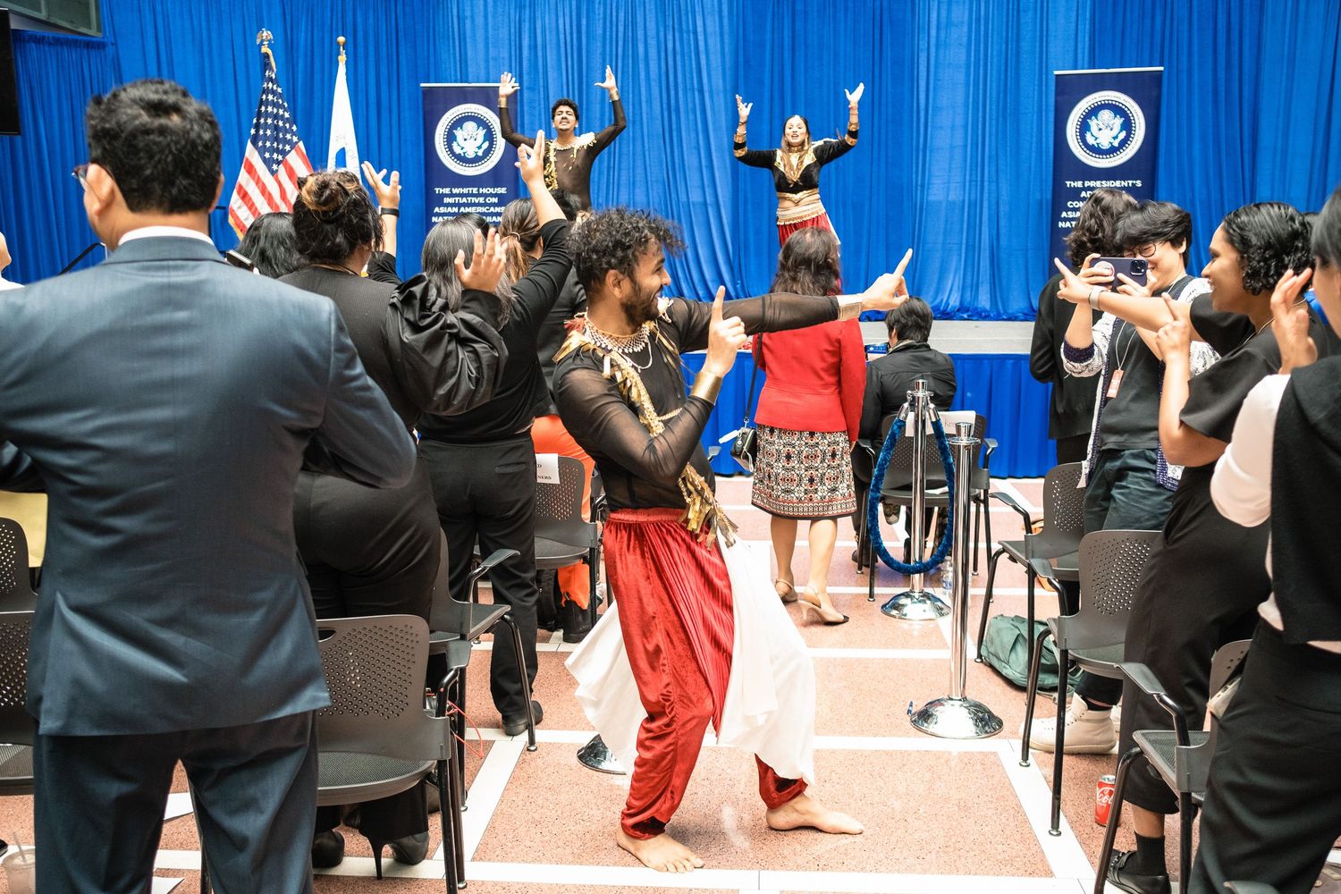 Performers in colorful traditional attire dance at a formal government office cultural celebration event.