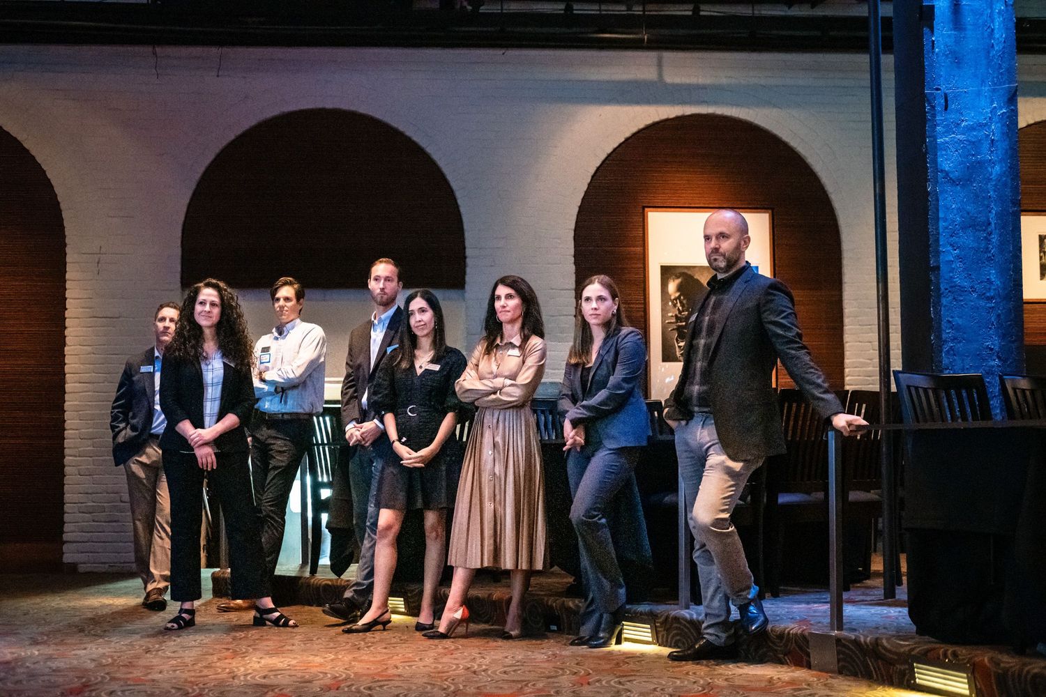 Group of seven professionals posing together on a stage set with arched windows and dramatic lighting.
