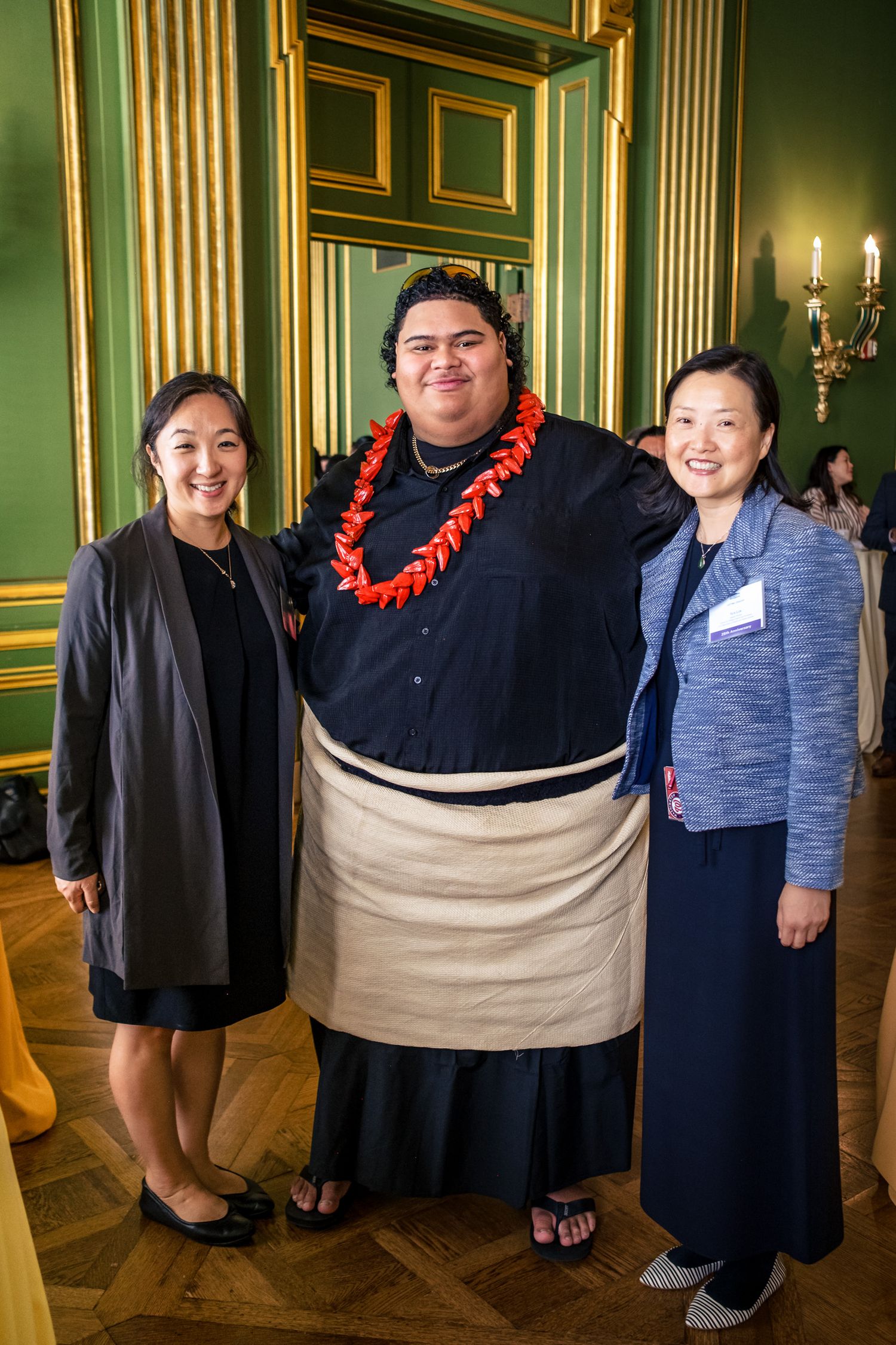 Two women pose with a large man in traditional sumo attire and red lei garland in an elegant green room.
