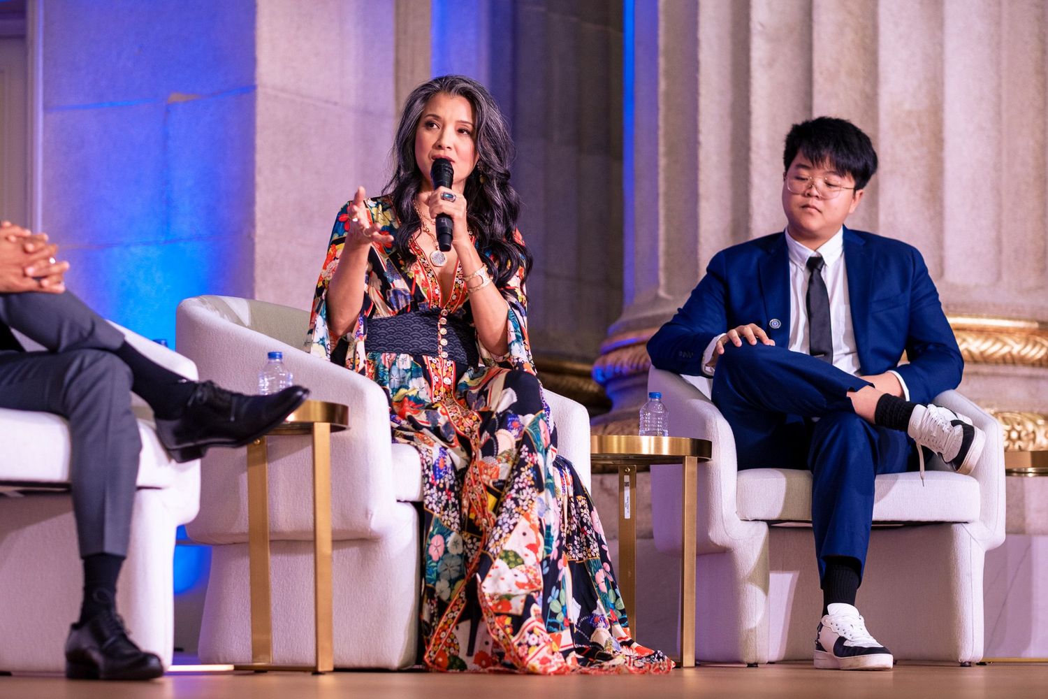 Woman in floral dress speaks at panel event flanked by two attendees on stage.